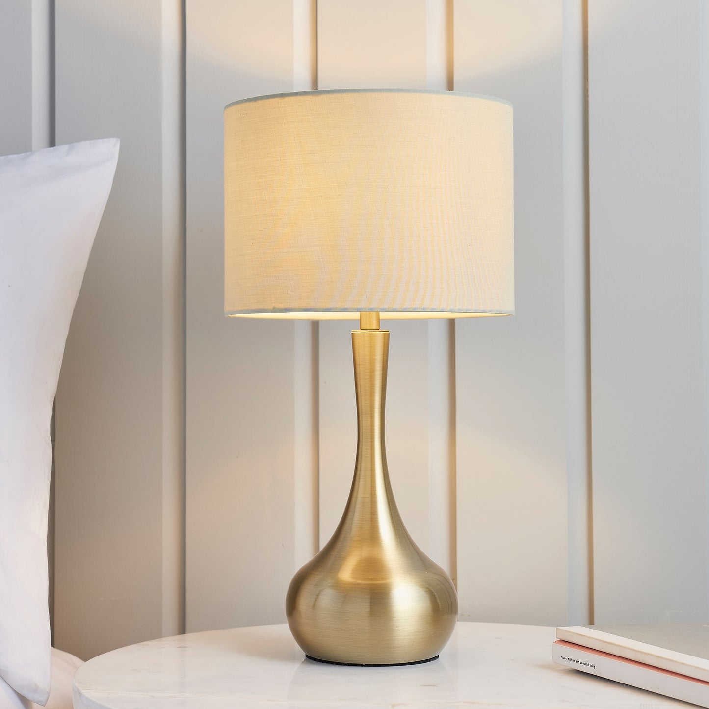 A brass and taupe Piccadilly table lamp from Kikiathome.co.uk enhances the interior decor of a white bed.