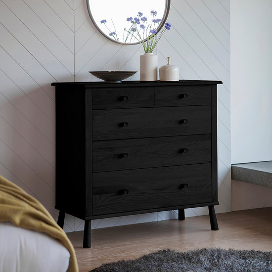 A Tigley 5 Drawer Chest Black 980x450x954mm in a bedroom with a mirror is stylish interior decor.