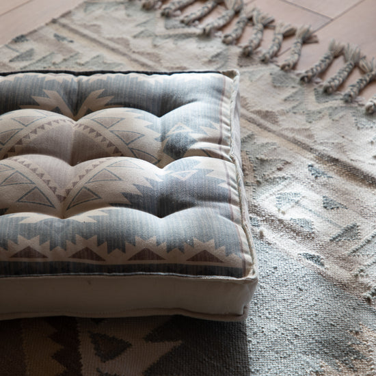 A grey floor cushion with Aztec print, placed on top of a rug for interior decor.