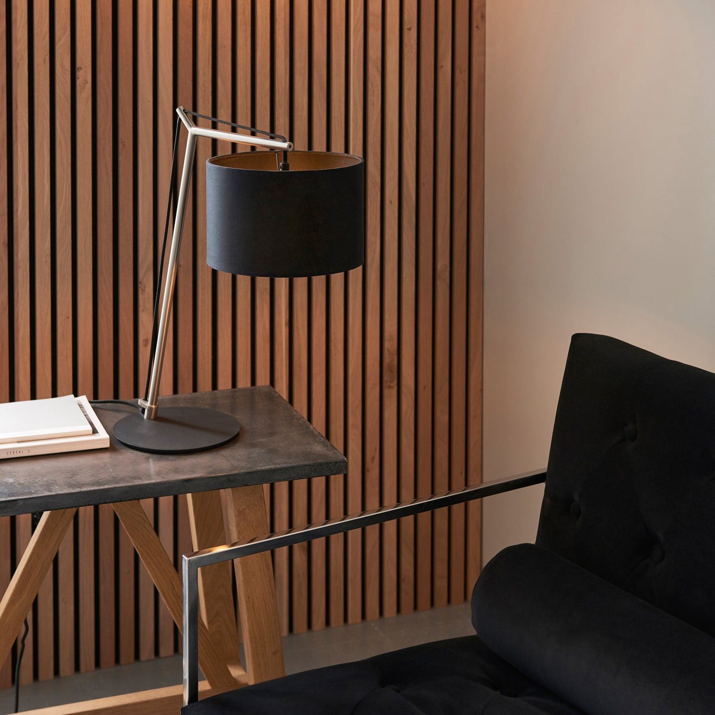 A black Buckland Table Lamp and antique Nickel chair and table in a room with wooden slats, perfect for home furniture and interior decor from Kikiathome.co.uk.