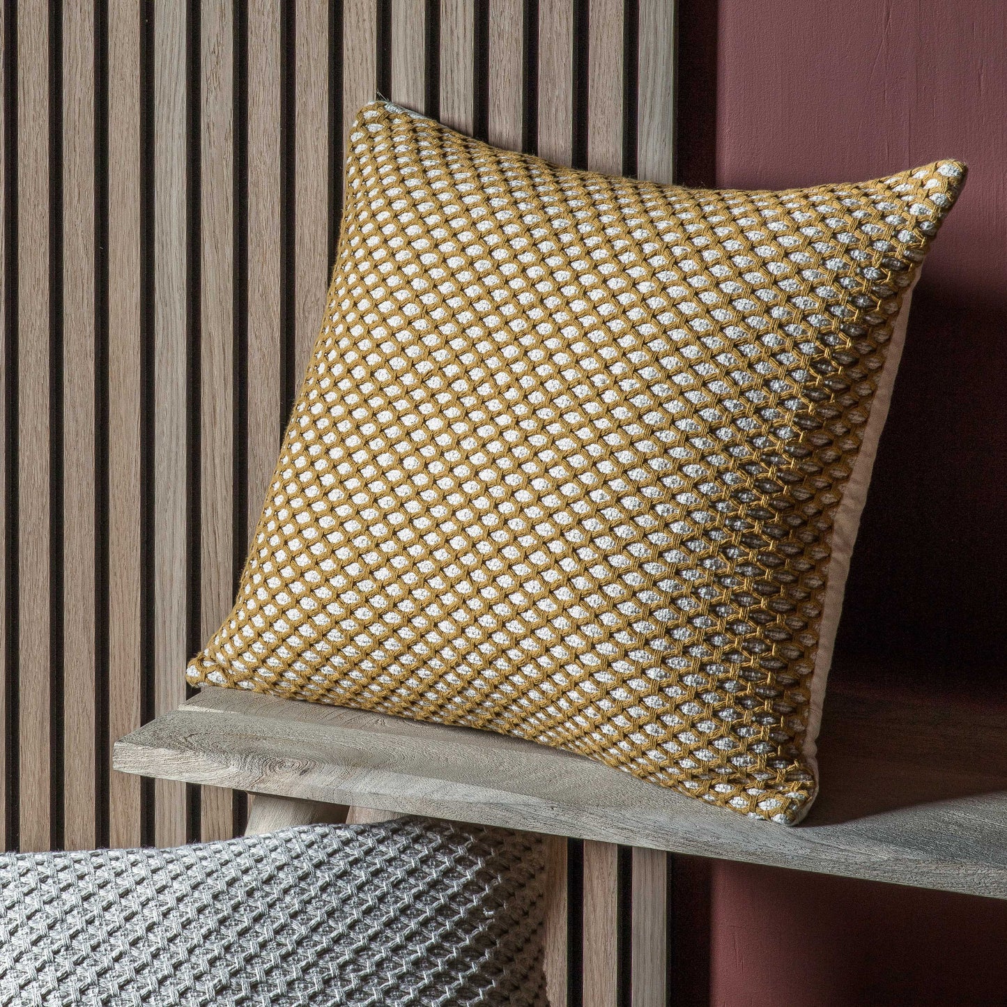 A Bittaford Cushion Ochre 550x550mm by Kikiathome.co.uk on a wooden shelf, perfect for interior decor.