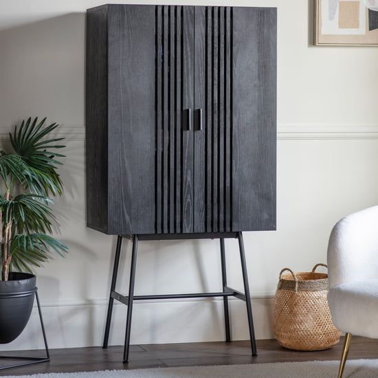 A stylish Holston Drinks Cabinet in Black complements the interior decor of a living room, accompanied by a potted plant for added home charm.