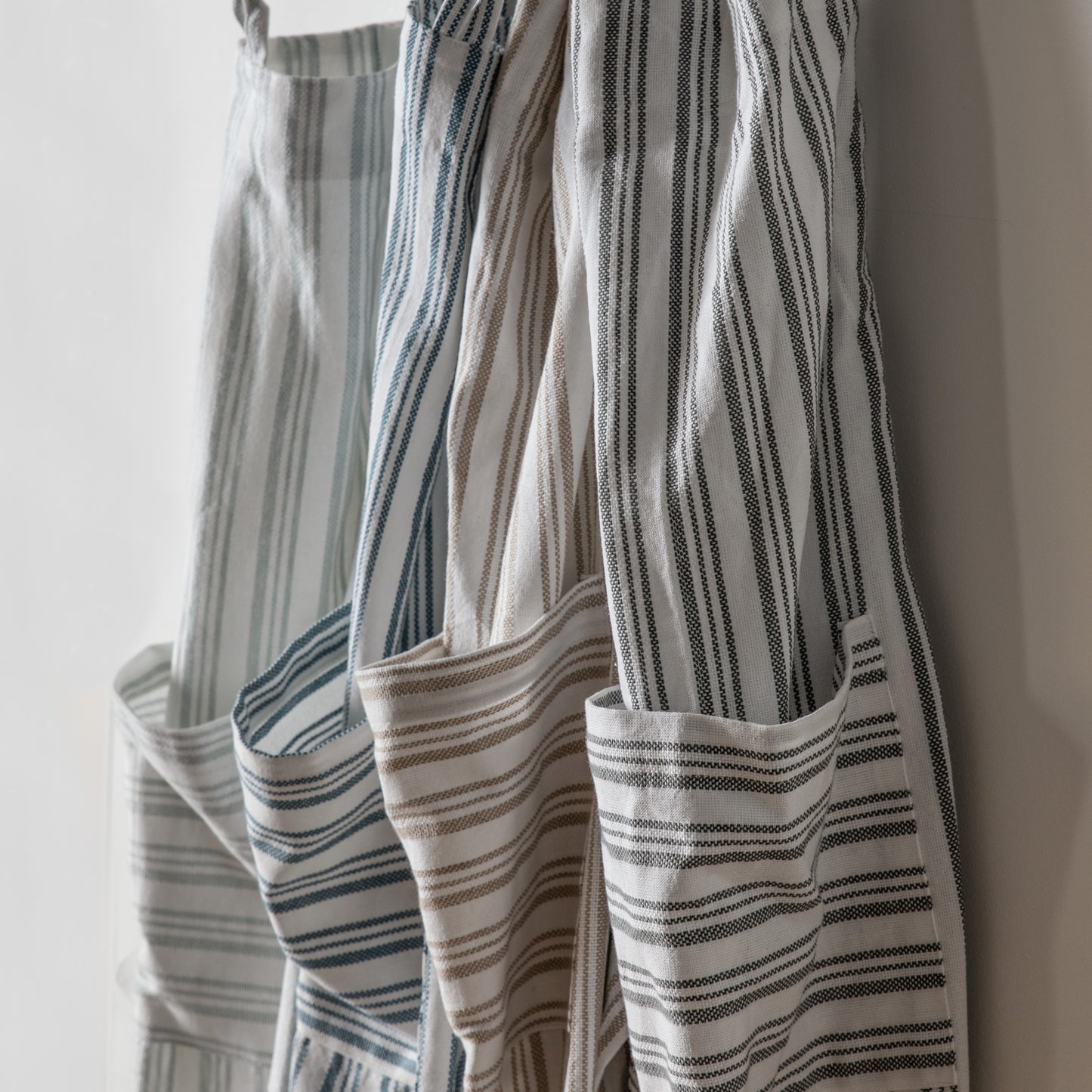 Load image into Gallery viewer, Three Organic Cotton Aprons hanging on a wall as part of the interior decor by Kikiathome.co.uk.
