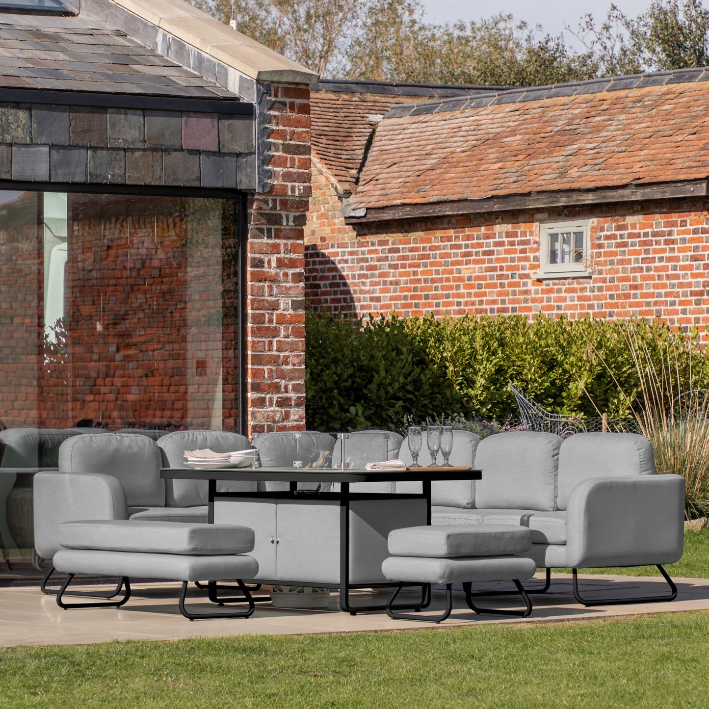 A patio with a Topsham Rect Dining Set with Fire Pit Table Slate from Kikiathome.co.uk, ideal for home furniture and interior decor, situated in front of a brick house.