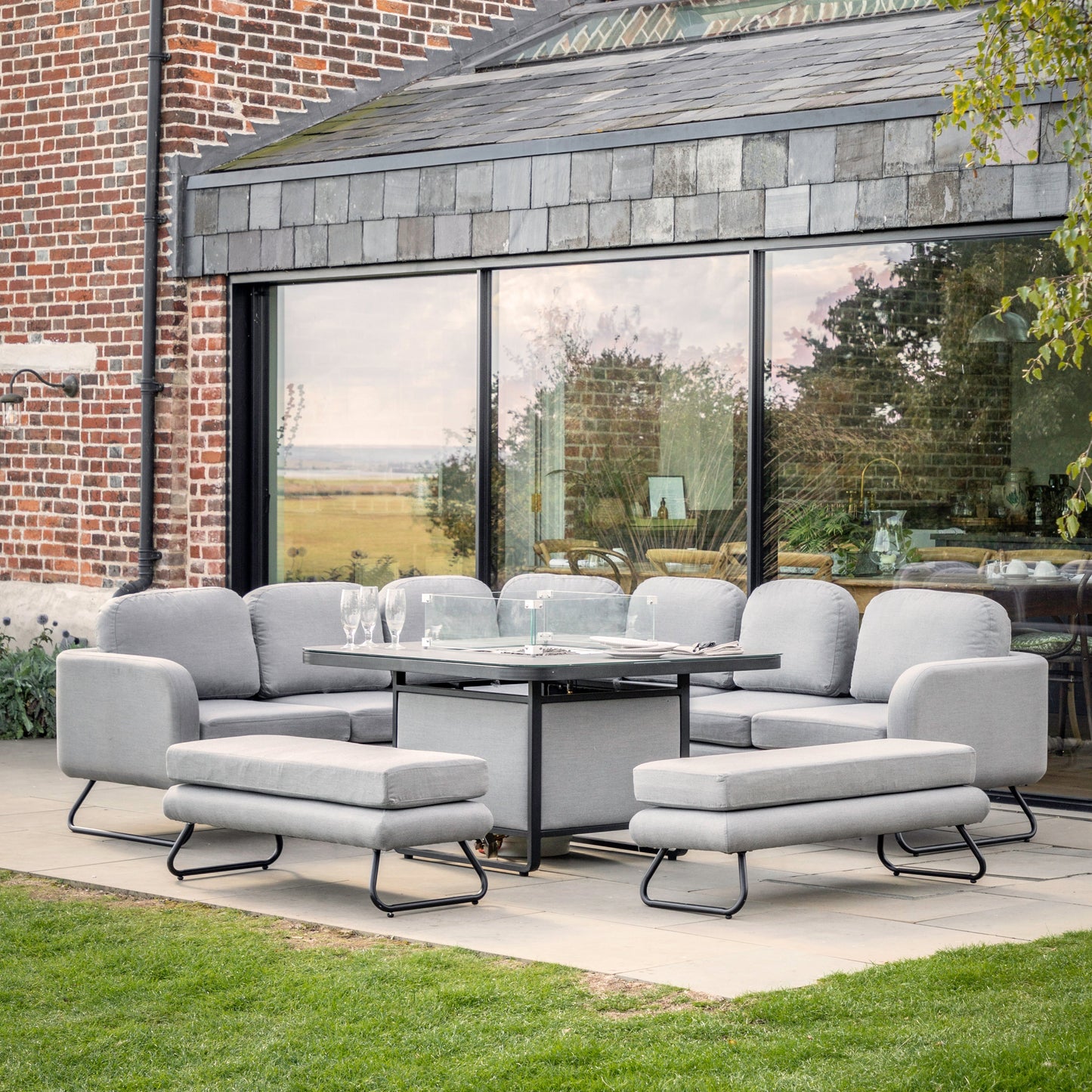 A sleek grey Topsham Dining Set with Fire Pit Table Slate from Kikiathome.co.uk enhances this glass house's interior decor.